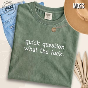 May include: A moss green crew neck t-shirt featuring the white text "quick question what the fuck." The shirt has a Comfort Colors label at the inner neckline. A gold-toned heart pendant necklace rests on the fabric near the collar.