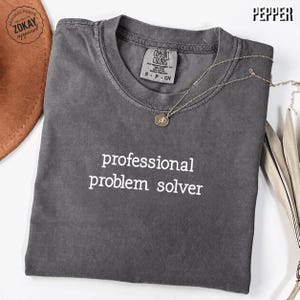 May include: A charcoal gray short-sleeve t-shirt with the white text "professional problem solver" printed on the chest. The shirt features a crew neckline and a Comfort Colors brand label on the inside collar. A thin gold-tone necklace rests on top.