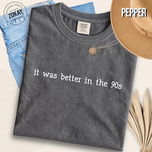 May include: A charcoal gray short-sleeve t-shirt featuring the white text "it was better in the 90s" printed across the chest. The shirt has a crew neckline and a visible brand label on the inside collar. The fabric has a washed, vintage appearance.
