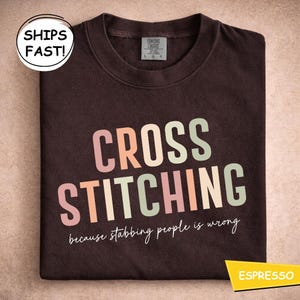May include: A dark brown t-shirt with the words "CROSS STITCHING" in pastel colours. The phrase "because stabbing people is wrong" is below the text. The shirt is folded and has a Comfort Colors label.