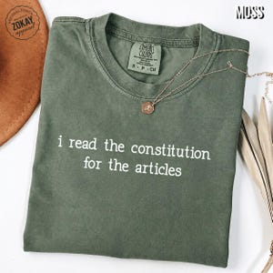 May include: A moss green short-sleeve t-shirt with a crew neckline and the white text "i read the constitution for the articles" printed on the front. The shirt features a Comfort Colors brand label at the inner neck and a delicate gold-tone necklace.