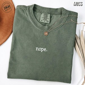 May include: A moss green short-sleeved crew neck t-shirt featuring the word nope. printed in white lowercase letters on the chest. The shirt has a Comfort Colors brand label inside the collar and is paired with a thin gold chain necklace with a small pendant.