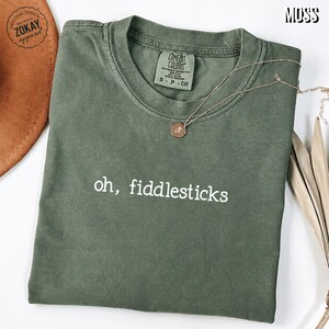 May include: A moss green crew neck t-shirt featuring the white text oh, fiddlesticks printed on the front. The shirt has a Comfort Colors label inside the collar and is displayed with a gold-toned necklace featuring a small circular pendant.