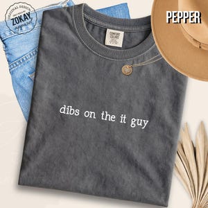 May include: A charcoal gray short-sleeve t-shirt with the white text "dibs on the it guy" printed on the front. The shirt features a crew neckline and a Comfort Colors brand label on the inside collar. The fabric has a washed, vintage appearance.