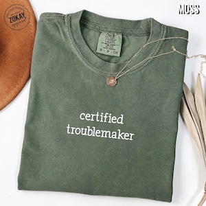 May include: A moss green crewneck t-shirt featuring the white text "certified troublemaker" printed on the chest. The shirt has a Comfort Colors brand label inside the collar and is displayed with a gold-toned necklace and a brown hat.
