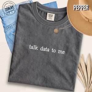 Peut inclure: Un t-shirt à manches courtes gris anthracite avec le texte blanc talk data to me imprimé sur le devant. Le t-shirt présente une encolure ronde et une étiquette de marque Comfort Colors à l'intérieur du col. Le tissu a un aspect délavé.