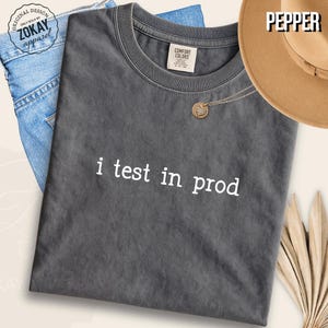 May include: A charcoal gray short-sleeve t-shirt with a crew neckline featuring the white text "i test in prod" printed on the front. The shirt has a faded, washed-out appearance and includes a Comfort Colors brand label at the inner neck.