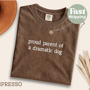 May include: Espresso-colored t-shirt with the text "proud parent of a dramatic dog" in white. A silver necklace with a heart-shaped pendant rests on the shirt. A brown hat is visible in the upper left corner.