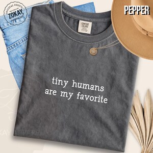 Peut inclure: Un t-shirt à col rond gris anthracite avec le texte blanc "tiny humans are my favorite" imprimé sur le devant. Le t-shirt présente une étiquette Comfort Colors à l'encolure intérieure et est présenté avec du tissu denim bleu et un chapeau beige.