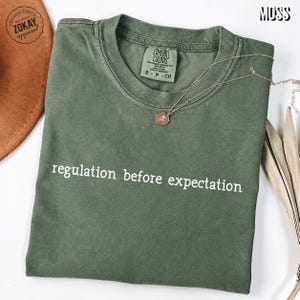 Peut inclure: Un t-shirt à col rond vert mousse avec le texte blanc "regulation before expectation" imprimé sur la poitrine. Le t-shirt porte une étiquette Comfort Colors à l'intérieur du col indiquant une taille small, accompagné d'un collier à chaîne dorée.