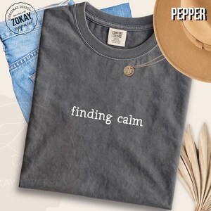 May include: A charcoal gray short-sleeve t-shirt with a crew neckline featuring the white text finding calm printed on the chest. The shirt has a vintage-washed appearance and includes a Comfort Colors brand label inside the collar area.