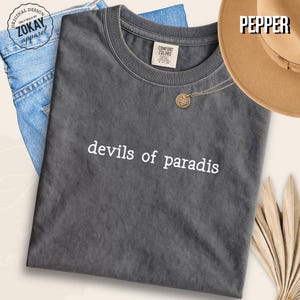 May include: A charcoal gray short-sleeve t-shirt with a crew neckline featuring the white text "devils of paradis" printed across the chest. The shirt has a visible brand label on the inside collar and a textured, washed-out fabric appearance.