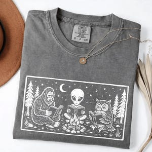 May include: A gray Comfort Colors t-shirt with a white graphic of a Bigfoot, an alien, and an owl reading books around a campfire under a crescent moon and stars. A gold necklace and a brown hat are also visible.