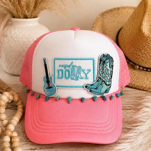 May include: A pink and white trucker hat featuring a white front panel with a light blue embroidered patch that reads "raised on DOLLY". The hat includes a blue guitar patch, a blue cowboy boot patch, and a string of small turquoise beads.
