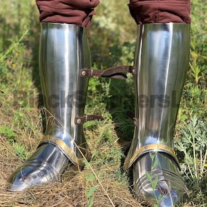 May include: Polished steel leg armor with brown leather straps and brass accents. The armor is highly reflective, showing the surrounding green grass and brown ground. The design is a historical recreation, likely for costume or display.