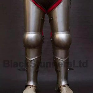 May include: Full leg armor made of polished metal, featuring articulated knee and shin plates. The armor is worn over black fabric with red trim. The image includes the text "BlackStunnersLtd".