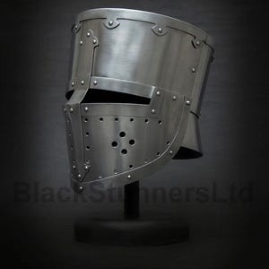 Op de afbeelding: Een zilverkleurige middeleeuwse helm met klinknagels en een vizier. De helm heeft een afgeronde bovenkant en een gezichtsbescherming met meerdere kleine gaten. De tekst "BlackStunnersLtd" is zichtbaar onderaan.