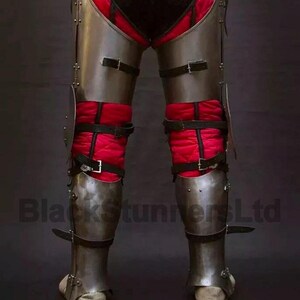 May include: Steel leg armor with black leather straps and red quilted padding. The armor is designed to protect the legs and feet. The image shows the lower body of a person wearing the armor. The text "BlackStunnersLtd" is visible.