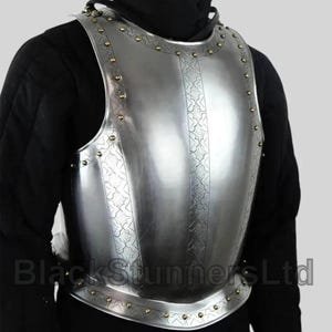 Peut inclure: Une cuirasse en acier poli avec des motifs gravés et des rivets en laiton sur les bords. L'armure est conçue pour protéger le torse et est portée sur un vêtement noir. Le texte "Black Stunners Ltd" est visible.