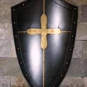 May include: A black shield with a gold cross design. The shield has a pointed bottom and is trimmed with gold rivets. The cross is centered on the shield and is made of gold-colored metal. The shield is mounted on a stone wall.