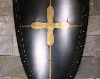 May include: A black shield with a gold cross design. The shield has a pointed bottom and is trimmed with gold rivets. The cross is centered on the shield and is made of gold-colored metal. The shield is mounted on a stone wall.