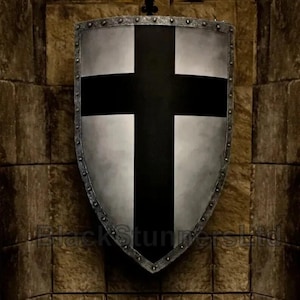 May include: A silver-finished shield with a black cross design, reminiscent of medieval heraldry. The shield features a riveted edge and is displayed against a textured stone wall, suggesting a historical or fantasy setting.