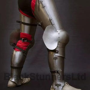 May include: Steel leg armor with articulated plates and knee protection. The armor features a polished metal finish, secured with leather straps and rivets. Red padded fabric is visible beneath the armor. The image shows a close-up of the armor.