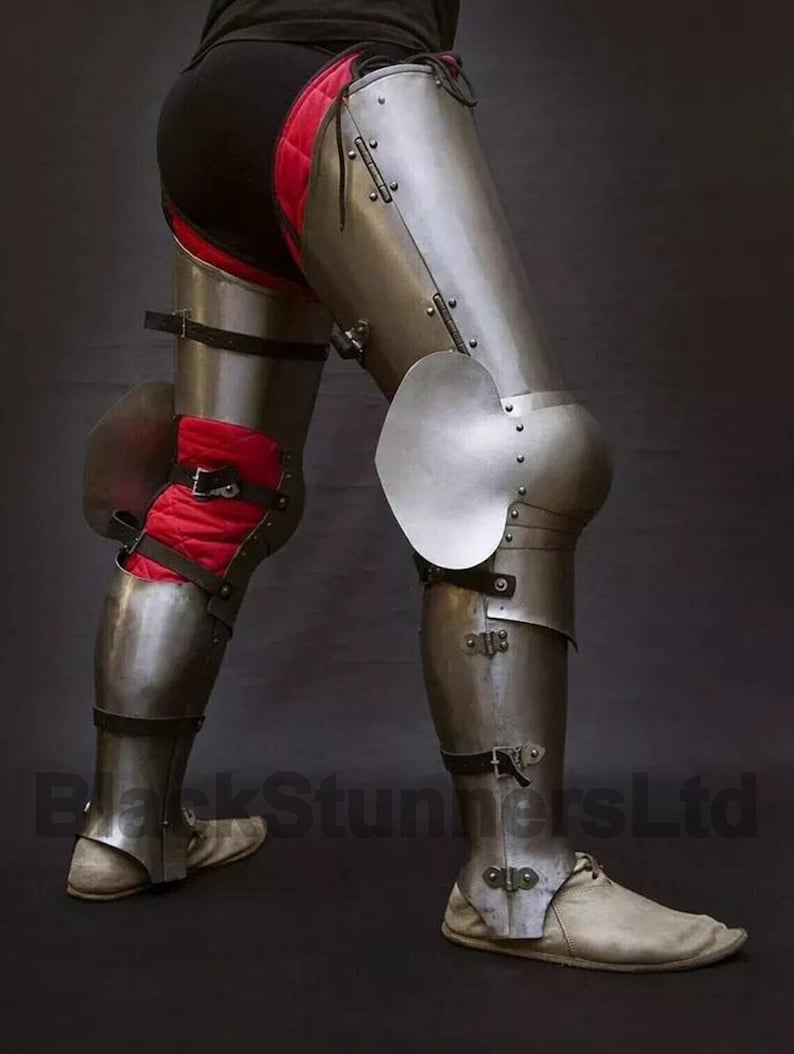 May include: Steel leg armor with articulated plates and knee protection. The armor features a polished metal finish, secured with leather straps and rivets. Red padded fabric is visible beneath the armor. The image shows a close-up of the armor.
