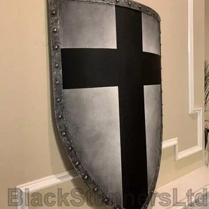 May include: A medieval-style shield with a silver and black design. The shield has a large black cross on a silver background, with a riveted silver border. The shield is mounted on a wall.