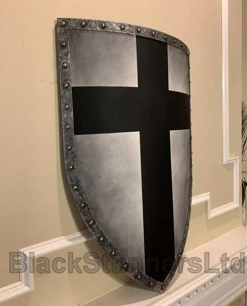 May include: A medieval-style shield with a silver and black design. The shield has a large black cross on a silver background, with a riveted silver border. The shield is mounted on a wall.
