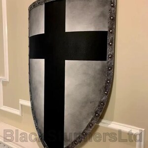 May include: A medieval-style shield with a black cross on a silver and gray background. The shield has a riveted, metallic border and is displayed against a neutral wall. The text "Black Sturmers Ltd" is visible at the bottom.