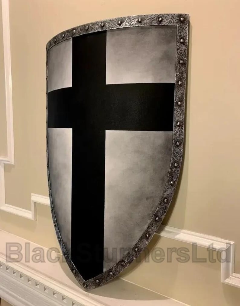 May include: A medieval-style shield with a black cross on a silver and gray background. The shield has a riveted, metallic border and is displayed against a neutral wall. The text "Black Sturmers Ltd" is visible at the bottom.
