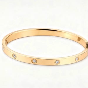 May include: A gold-coloured bangle bracelet with six small, round, clear gemstones evenly spaced around the band. The bracelet has a smooth, polished finish and a hinged clasp. The background is plain white.