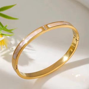 May include: Gold-coloured bangle bracelet featuring a white mother-of-pearl inlay and a small diamond accent. The bracelet has a hinged clasp. A stylish accessory.