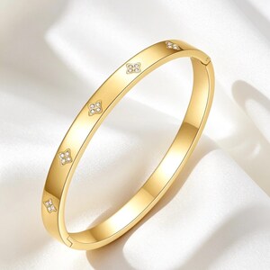 May include: A gold-coloured bangle bracelet with a clasp closure. The bracelet features four decorative floral designs, each with four small, clear gemstones. The bracelet is displayed on a white, silky surface.