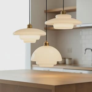 May include: Three pendant lights with a layered, off-white glass shade and brass-colored hardware. The lights hang from black cords, illuminating a kitchen setting. The design features a tiered, saucer-like shape, offering a modern aesthetic.