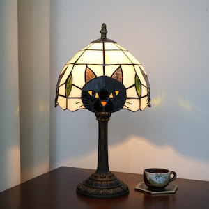Op de afbeelding: Een gebrandschilderde tafellamp met een zwart kattenontwerp. De lamp heeft een crèmekleurige kap met groene en bruine accenten en een zwart kattengezicht in het midden. De voet is donkerbrons en er staat een kleine theekop in de buurt.