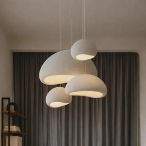 Op de afbeelding: Vier moderne, crèmekleurige hanglampen van verschillende formaten hangen aan het plafond. Elke lamp heeft een afgeronde, organische vorm met een zacht, getextureerd oppervlak en een warme, gloeiende binnenkant. De lampen hangen tegen een achtergrond van grijze gordijnen.