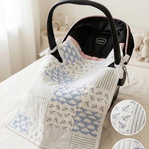 May include: A baby car seat with a custom-made patchwork cover. The cover features a mix of white, blue, and gray fabrics with cloud and paper airplane patterns. The car seat is black with a pink trim and a black handle. The cover is tied to the car seat with white ribbons. The text "HANDMADE WITH LOVE" is visible.