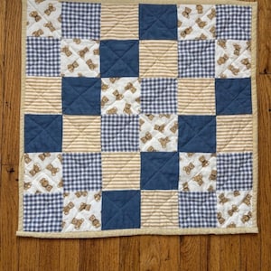May include: A patchwork quilt with a beige border. The quilt features squares of navy blue, beige stripes, white with brown teddy bears, and blue and white gingham. The text "Made By Summer Studio" is at the bottom.