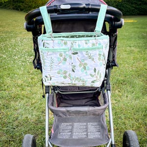 May include: A stroller organizer bag with a white background and a green leaf pattern. The bag has two pockets and mint green straps. The stroller is black and gray with a pink accent.