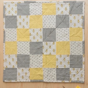 May include: A patchwork quilt with a repeating pattern of yellow, gray, and white squares. Some squares feature duckling motifs, stripes, or polka dots. The quilt is surrounded by a wooden surface, with sewing tools and the text "Made By Summer Studio".