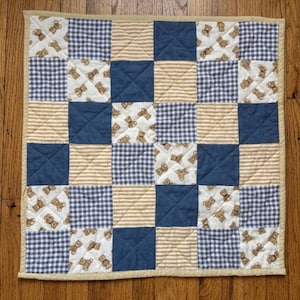 May include: A square patchwork quilt featuring a repeating pattern of teddy bears on a white background, alternating with solid navy blue squares, tan and white striped squares, and navy and white gingham checkered squares with a tan border.
