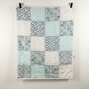 May include: A patchwork baby quilt with a white border, featuring squares of light blue, white with gray clouds, white with gray stars, and gray elephants. The quilt is hanging from black clips against a white wall.