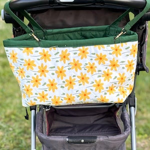 May include: A stroller organizer bag with a white background and a pattern of yellow daisies. The bag has green trim and straps, and is attached to a stroller. The text "Made By Summer Studio" is visible at the bottom.