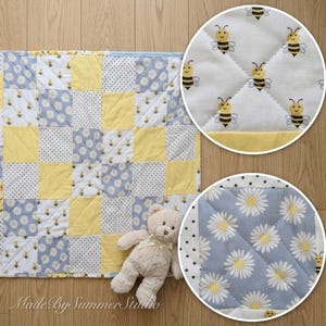 May include: A patchwork baby quilt with a mix of yellow, white, and blue squares. Some squares feature daisy and bee patterns. A small teddy bear rests on the quilt. The text "Made By Summer Studio" is visible.