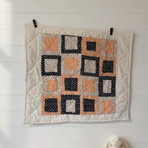 May include: A square quilt with a patchwork design of peach, navy, and white squares. The quilt is hanging on a white wall with black clips. A stuffed bunny and wooden blocks are in the foreground.