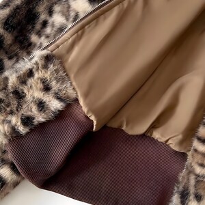 May include: A close-up of a faux fur jacket with a leopard print pattern. The jacket features a brown ribbed cuff and a tan interior lining. A silver zipper runs down the front of the jacket.