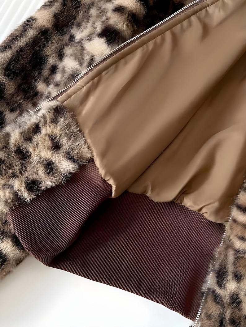 May include: A close-up of a faux fur jacket with a leopard print pattern. The jacket features a brown ribbed cuff and a tan interior lining. A silver zipper runs down the front of the jacket.