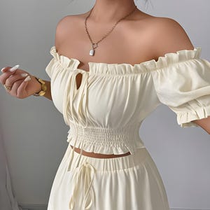 May include: A cream-colored two-piece set featuring an off-the-shoulder crop top with short ruffled sleeves, a smocked waist, and a center tie detail. The matching skirt includes a drawstring waist and a horizontal lace trim detail near the hem.
